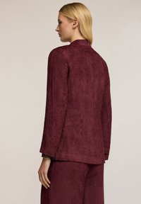 Blazer testurizzato bordeaux con maniche lunghe e colletto rialzato, caratterizzato da una silhouette dritta e dettagli minimi.