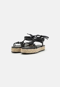 Sandali neri intrecciati con cinturini annodati, suole in juta intrecciata e una soletta piatta nera opaca. Design casual e a basso profilo.