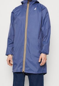 Blå lang regnfrakke med hætte, flerfarvet stribet lynlås, elastiske manchetter, sidelommer, båret over mørkeblå jeans.