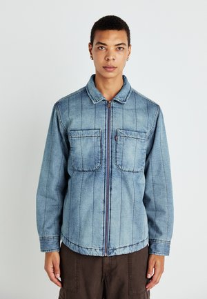 Chaqueta de mezclilla azul claro con cremallera frontal, dos bolsillos en el pecho y detalles de costura horizontal. Presenta un cuello con solapa y un dobladillo elástico.
