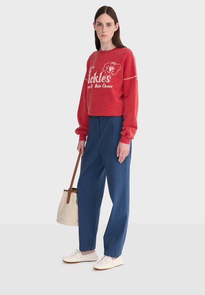 Kobieta w czerwonej bluzie z nadrukiem, niebieskich spodniach z wysokim stanem, białych butach, trzymająca beżową torebkę z brązowym paskiem, stojąca na jednolitym tle.