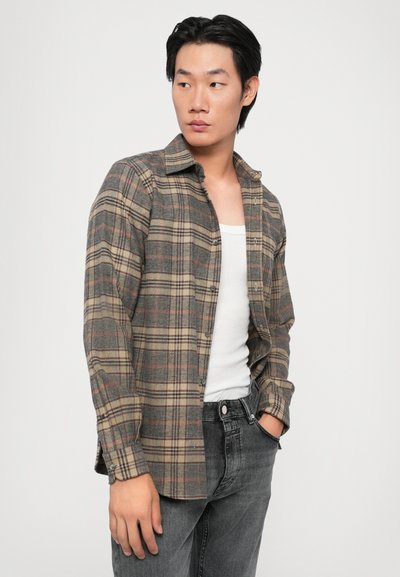 Chemise en flanelle à carreaux gris et beige, à manches longues, avec des poignets boutonnés, associée à un débardeur blanc et à un jean foncé.