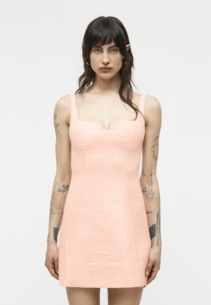 Kobieta o ciemnych włosach, ubrana w różową, bezrękawną mini sukienkę o fakturze, stojąca na tle jasnego, jednolitego tła, z widocznymi tatuażami na ramionach.