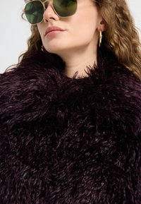 Kvinde med grønne solbriller, guldhoops øreringe og en mørk lilla, fluffy tekstureret frakke, vist fra halsen og op.