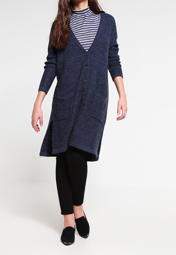 Femme portant un long cardigan bleu marine à boutons, un col roulé rayé, des leggings noirs et des chaussures noires sans lacets marchant vers l'avant.