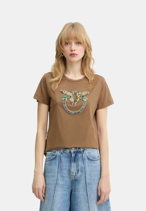 Blondína v hnedom tričku s farebným dizajnom vtáka z korálok a svetlomodrých džínsach s vysokým pásom, stojaca na jednoduchom pozadí.