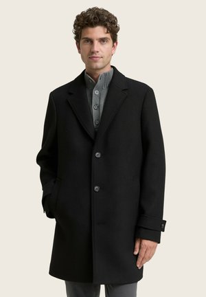 Black wool coat with notched lapels, three buttons, and side pockets. Worn over a gray ribbed sweater. Smooth texture, tailored fit.