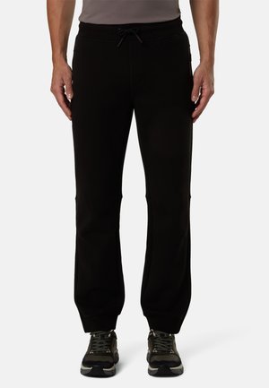 Uomo con pantaloni della tuta neri con coulisse e scarpe da ginnastica nere e grigie, in piedi davanti a uno sfondo semplice.