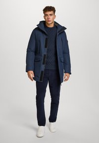 Un giovane in piedi indossa un parka imbottito blu navy, un maglione a trecce, pantaloni scuri e sneakers bianche su uno sfondo grigio chiaro.