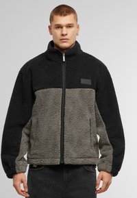 Fleece striukė su juodu viršumi ir šviesiai pilku apačiu, su užtrauktuku, aukštu apykakle ir šoninėmis kišenėmis. Tekstūruota medžiaga.