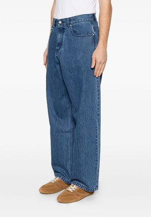 Persona che indossa jeans blu larghi in denim con chiusura con bottone e zip, abbinati a scarpe stringate in camoscio marrone.