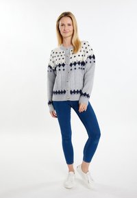 Cardigan grigio lavorato a maglia con motivi geometrici bianchi e blu navy, dotato di scollatura a girocollo, chiusura con bottoni e polsini a costine, abbinato a jeans blu e sneakers bianche.