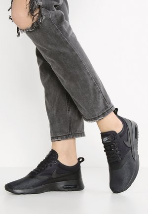 Osoba nosząca czarne sneakersy Nike i podarte ciemnoszare dżinsy, stojąca na białym tle.