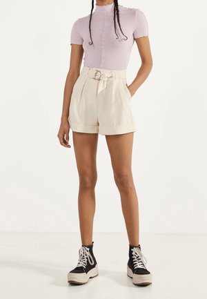 Top aderente viola chiaro con maniche corte e scollo smerlato, abbinato a pantaloncini crema a vita alta con cintura e tasche.