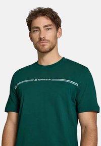 Temnozeleni bombažni majica s kratkimi rokavi, okroglim izrezom in belimi črtami sLOGO-jem čez prsni del. Gladka tekstura, sproščen dizajn.