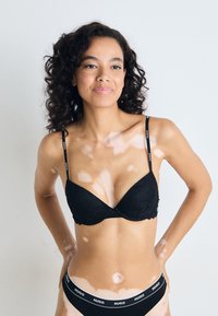 Black lace bra with thin straps featuring a logo, paired with matching black brief adorned with a logo waistband. Smooth texture, fitted design.
