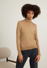 Világosbarna garbós pulóver puha kötött anyagból, slim fit fazonnal, hosszú ujjakkal és oldalsó hasítékokkal az alján. Sötét farmernadrággal párosítva.