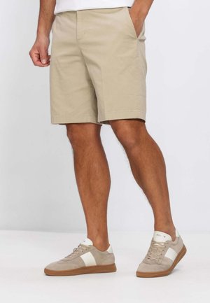 Uomo che indossa pantaloncini beige e scarpe da ginnastica beige e bianche, in piedi con una mano in tasca contro uno sfondo uniforme.