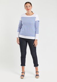 Witte langärmelige top met blauwe horizontale strepen, gecombineerd met donkere jeans. Het model draagt beige platform sandalen en laat een ontspannen pasvorm zien.