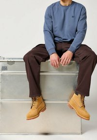 Blå sweatshirt med långa ärmar och en liten logotyp, bruna löst sittande byxor och gula stövlar med snören och gummisålar. Sitter på en silverlåda.