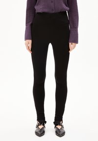 Zwarte leggings met een gladde textuur en een aansluitend ontwerp, gecombineerd met een gestructureerd paarse blouse met knopen en zwarte schoenen met enkelbandjes.