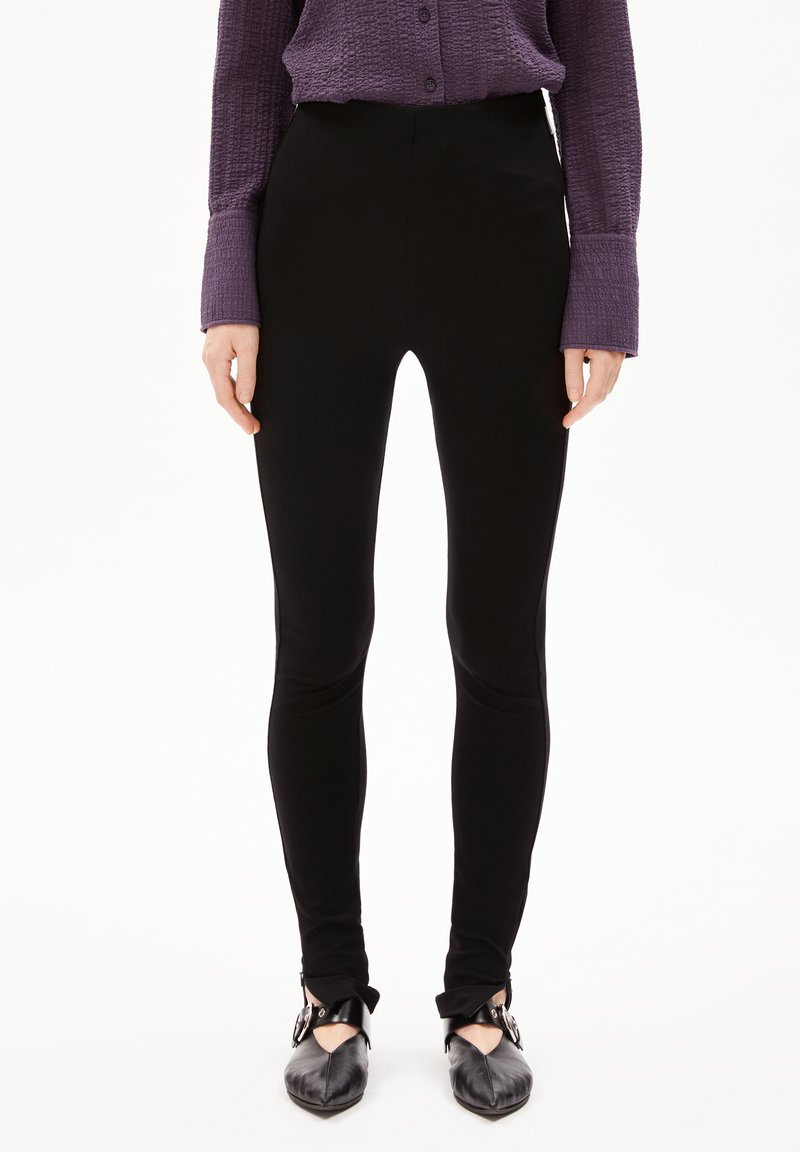 Zwarte leggings met een gladde textuur en een aansluitend ontwerp, gecombineerd met een gestructureerd paarse blouse met knopen en zwarte schoenen met enkelbandjes.