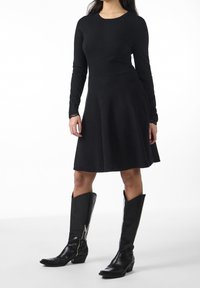 Robe noire à manches longues avec un corsage ajusté et une jupe évasée. Associée à des bottes en cuir noir montantes avec une fermeture éclair sur le côté.