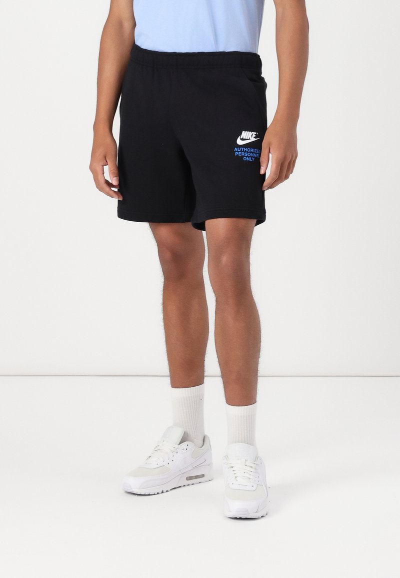 Juodi medvilniniai šortai su gumine juosta, ant kurių yra balta „Nike“ logo ir mėlynas tekstas „TIK ĮGALIOTAS PERSONNELIS“. Dėvimi su baltais sportbačiais.