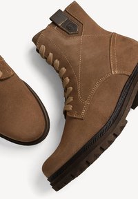 Stivali alla caviglia in suede marrone con design stringato, tacco rinforzato e suola in gomma testurizzata. Presentano un accento marrone in cima per facilitarne l'indossamento.