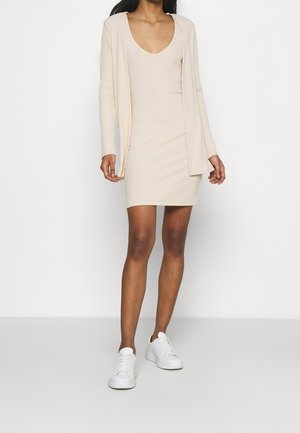 Kobieta ubrana w beżową, prążkowaną dzianinową sukienkę z dopasowanym, otwartym kardiganem i białe sneakersy, stojąca na jednolitym białym tle.