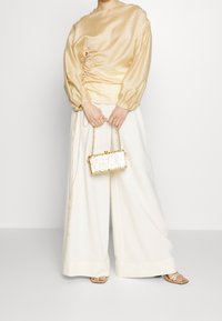 Satiinisestä kultaisesta paidasta, jossa on rypytetty yksityiskohta, on yhdistetty leveisiin valkoisiin housuihin sekä teksturoituun valkoiseen käsilaukkuun, jossa on kultaisia yksityiskohtia.