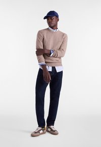 Beige cable-knit sweater over a light blue collared shirt, dark denim jeans, and beige sneakers with gray accents and white laces. Navy cap.