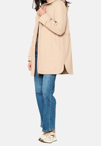 Beżowy długi płaszcz z kapturem, wykonany z miękkiego, teksturowanego materiału; posiada przednią kieszeń i luźny krój, noszony na niebieskie dżinsy i sneakersy.