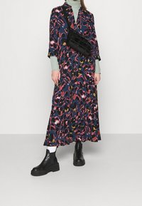 Lange jurk met een donkerblauwe achtergrond met een kleurrijke dierenprint in roze, geel en zwart; gecombineerd met zwarte chunky laarzen en een zwarte tas.
