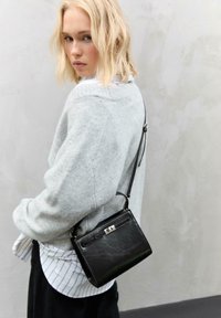 Sorth læderhåndtaske med en sølvlås, rektangulær form, båret som crossbody. Blød grå trøje over en hvidstribet skjorte.