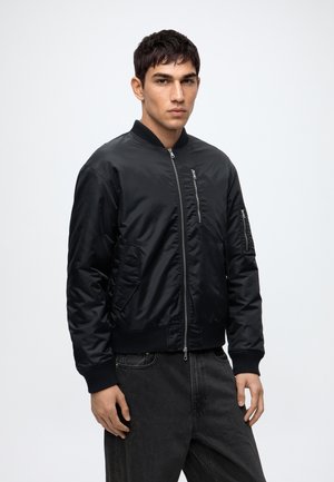 Giovane uomo con capelli corti e scuri che indossa una giacca bomber nera con cerniera e jeans scuri, in piedi davanti a uno sfondo chiaro e uniforme.