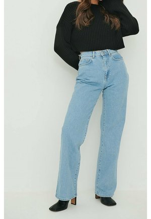 Femme portant un jean bleu clair taille haute à jambes larges, un pull noir court côtelé et des bottines noires à talons, sur un fond uni.
