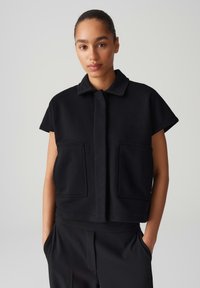 Schwarze, kurz geschnittene Jacke aus einem glatten Stoff, mit kurzen Ärmeln, einem Kragen und zwei aufgenähten Taschen vorne. Einfaches, klares Design.