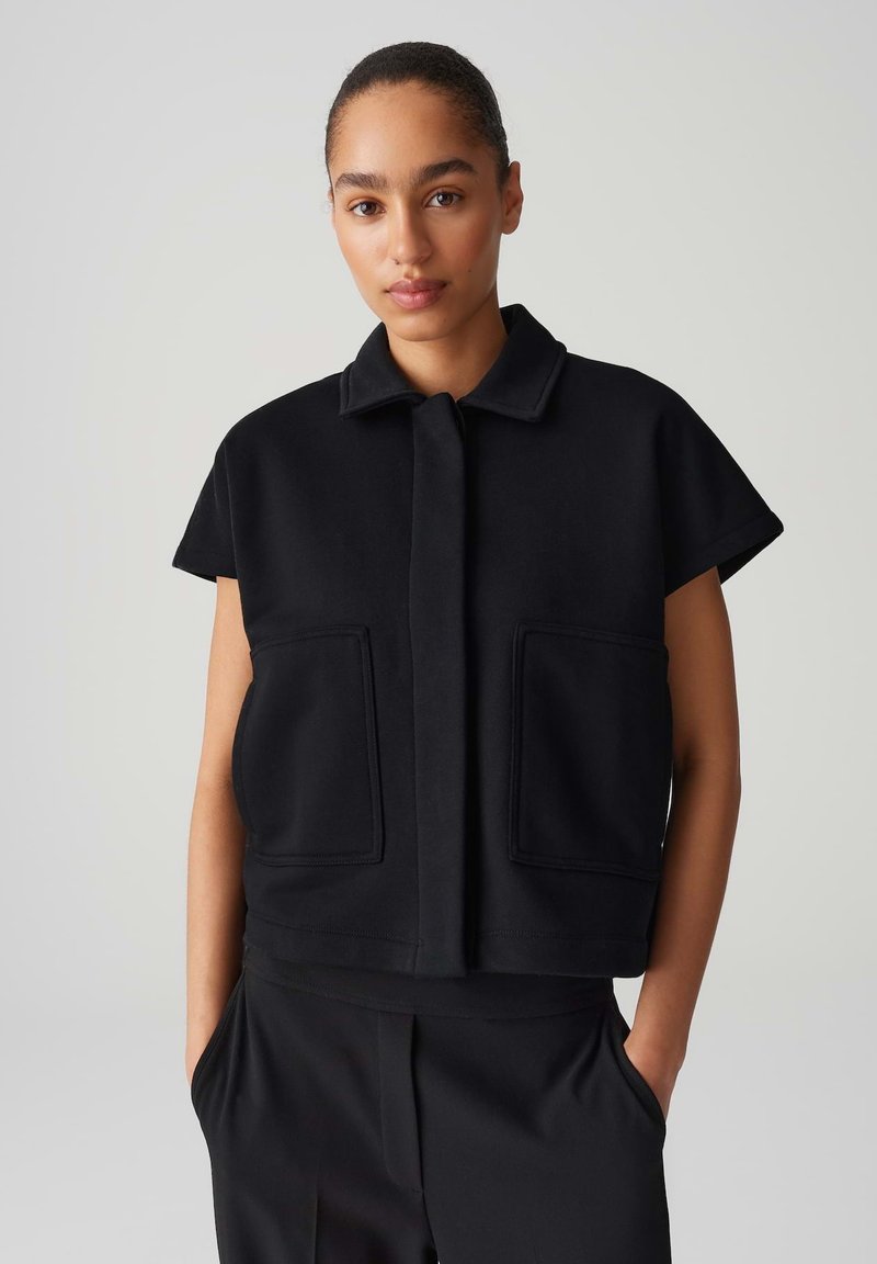 Schwarze, kurz geschnittene Jacke aus einem glatten Stoff, mit kurzen Ärmeln, einem Kragen und zwei aufgenähten Taschen vorne. Einfaches, klares Design.