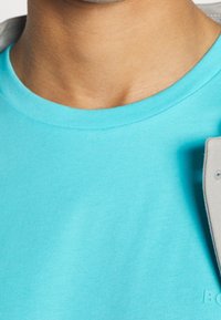 Tyrkysové bavlněné tričko s kulatým výstřihem. Má hladkou texturu a malou logo aplikaci na spodním lemu, v kombinaci se šedým límcem bundy.