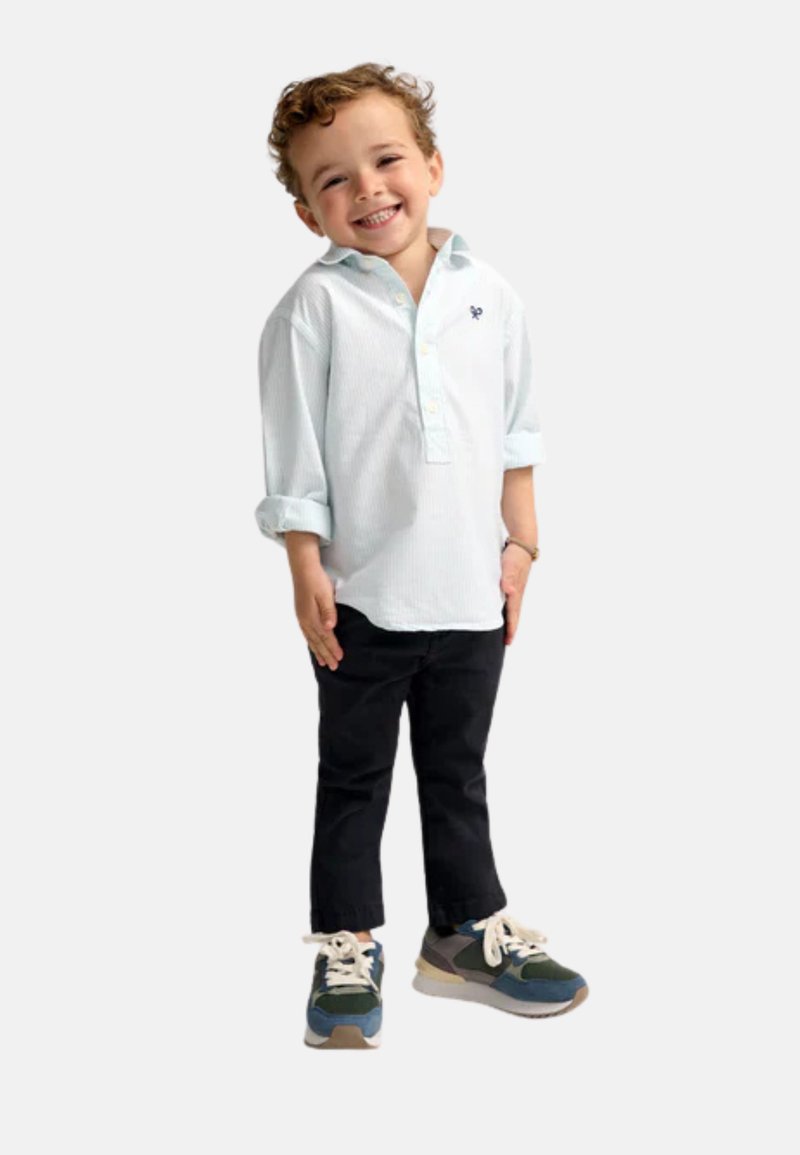 Camisa de botones a rayas azul clara, pantalones negros y zapatillas multicolores. Niño de pie con las manos en las caderas, sonriendo.