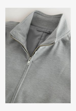 Veste zippée grise avec un col montant, texture lisse et détails en zip argenté. Le tissu semble doux et décontracté.