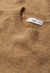 Beżowy sweter z dzianiny, z prążkowanym okrągłym dekoltem i teksturowanym materiałem. Widoczna metka z nazwą marki "MNG" i instrukcjami pielęgnacji.