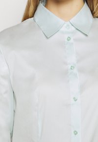 Chemise en satin bleu clair avec un col pointu et cinq boutons assortis sur le devant. Texture lisse sans motifs ni accents visibles.