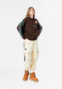 Joven con las manos en los bolsillos, vistiendo un sudadera marrón con mangas de texto azul, pantalones cargo color crema y botas de cordones color tostado.