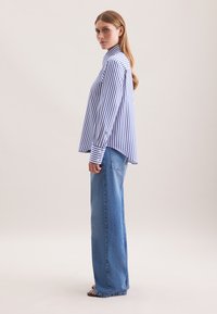 Vrouw die zijwaarts staat en een blauw-witte verticaal gestreepte blouse met lange mouwen en losse blauwe jeans draagt, met zwarte sandalen.