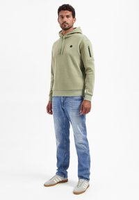 Lichtgroene hoodie met een voorzak in kangarustijl en een zijrits zak, gecombineerd met vervaagde blauwe jeans en grijze sneakers met witte strepen.