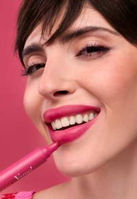 Marcador de labios rosa sostenido cerca de los labios, aplicando un tono rosa mate. Sonrisa brillante con dientes blancos, sobre un fondo rosa.