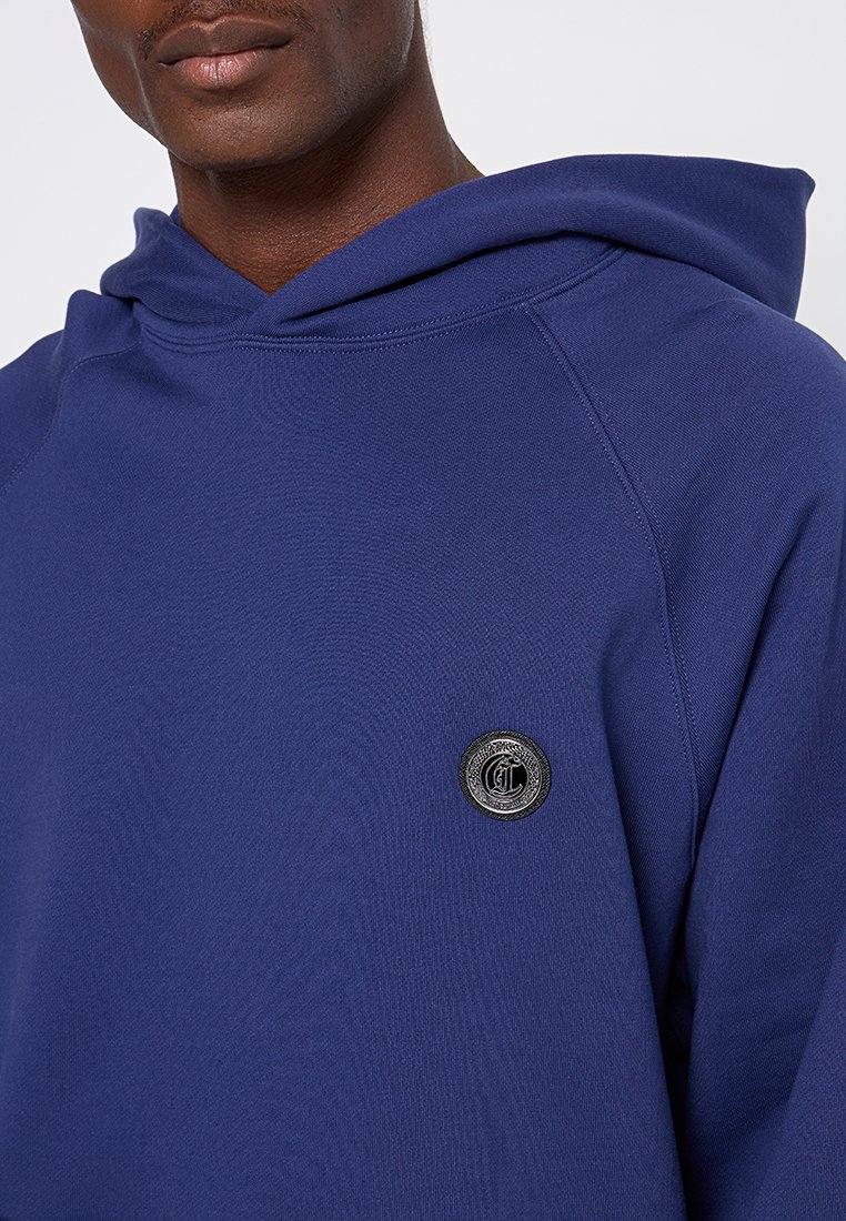 Granatowy hoodie o miękkiej fakturze, z okrągłym czarnym logo na klatce piersiowej oraz szerokim, strukturalnym kapturem.