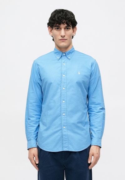 Chemise à manches longues bleu clair en coton, avec un col boutonné et un logo brodé discret sur la poitrine. Texture lisse.